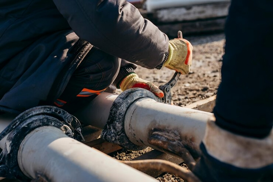 Two workers in protective gloves and clothing repair large metal pipes outdoors with hand tools, possibly for a burst pipe.