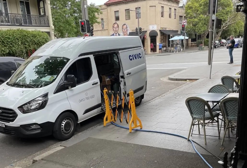 White civic van parked on city street with hoses attached and yellow barriers beside outdoor café seating.