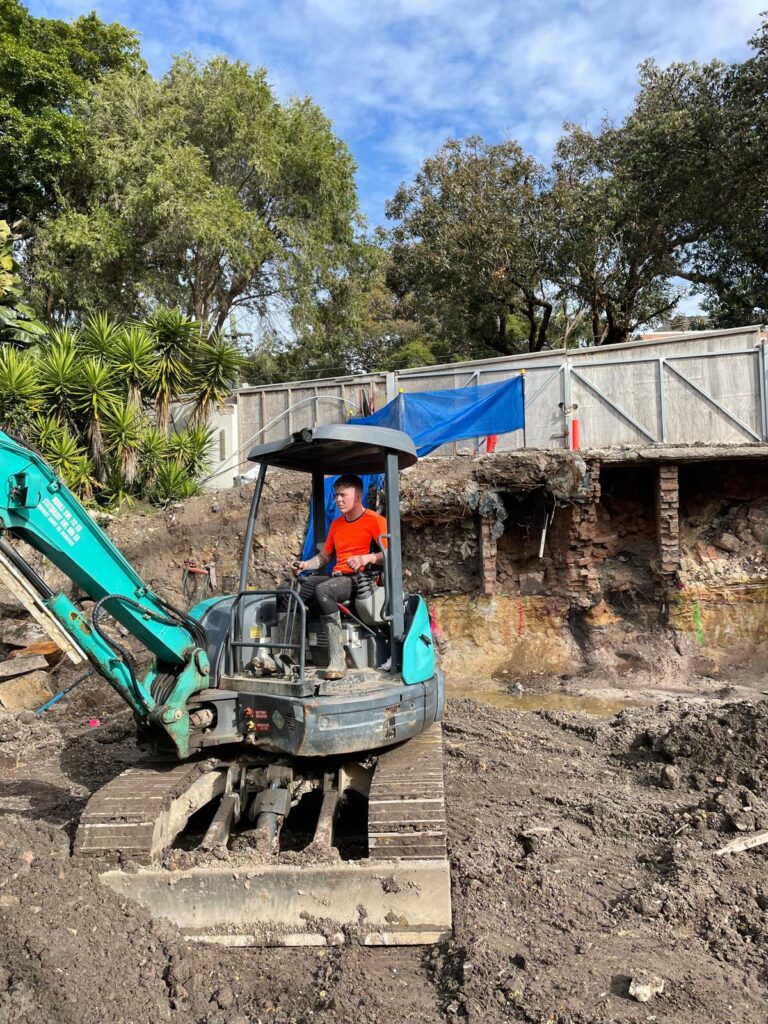 A Plumber in Eveleigh operates a small excavator on a site with dirt, trees, and a wooden fence in the background.