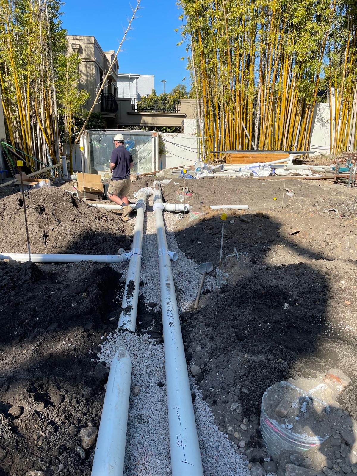 A Plumber in Randwick installs white PVC pipes in a trench at a construction site, with bamboo and buildings nearby.
