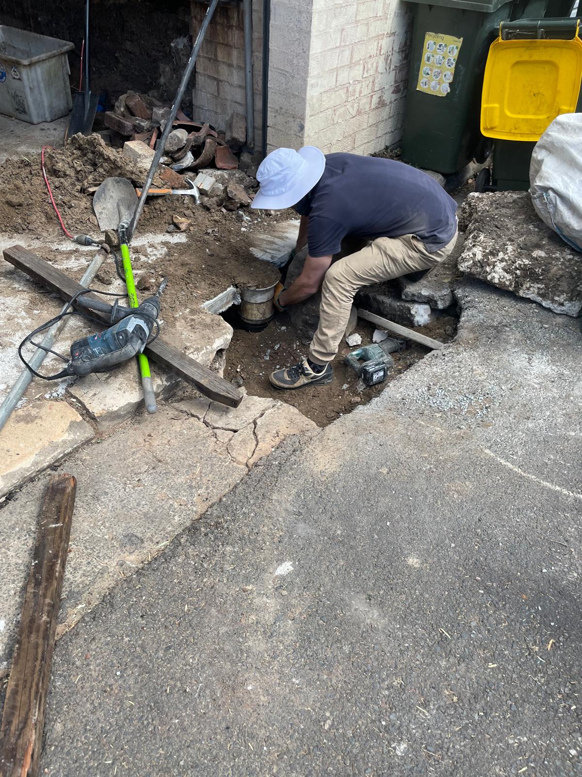 Redfern plumber kneeling in a dug-out by a driveway, working on a pipe with tools scattered, emergency service. 