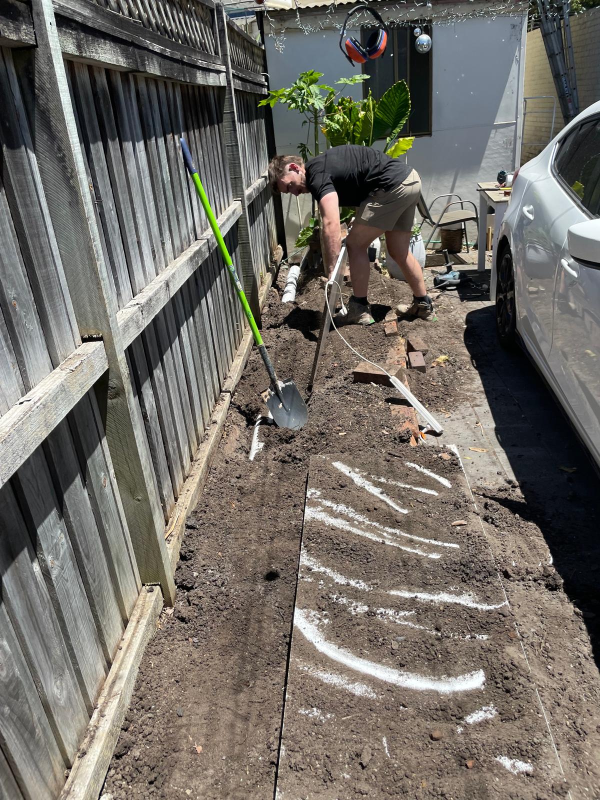A person digs soil by a wooden fence with a shovel; nearby are gardening tools, a car, and plumber Rosebery materials.