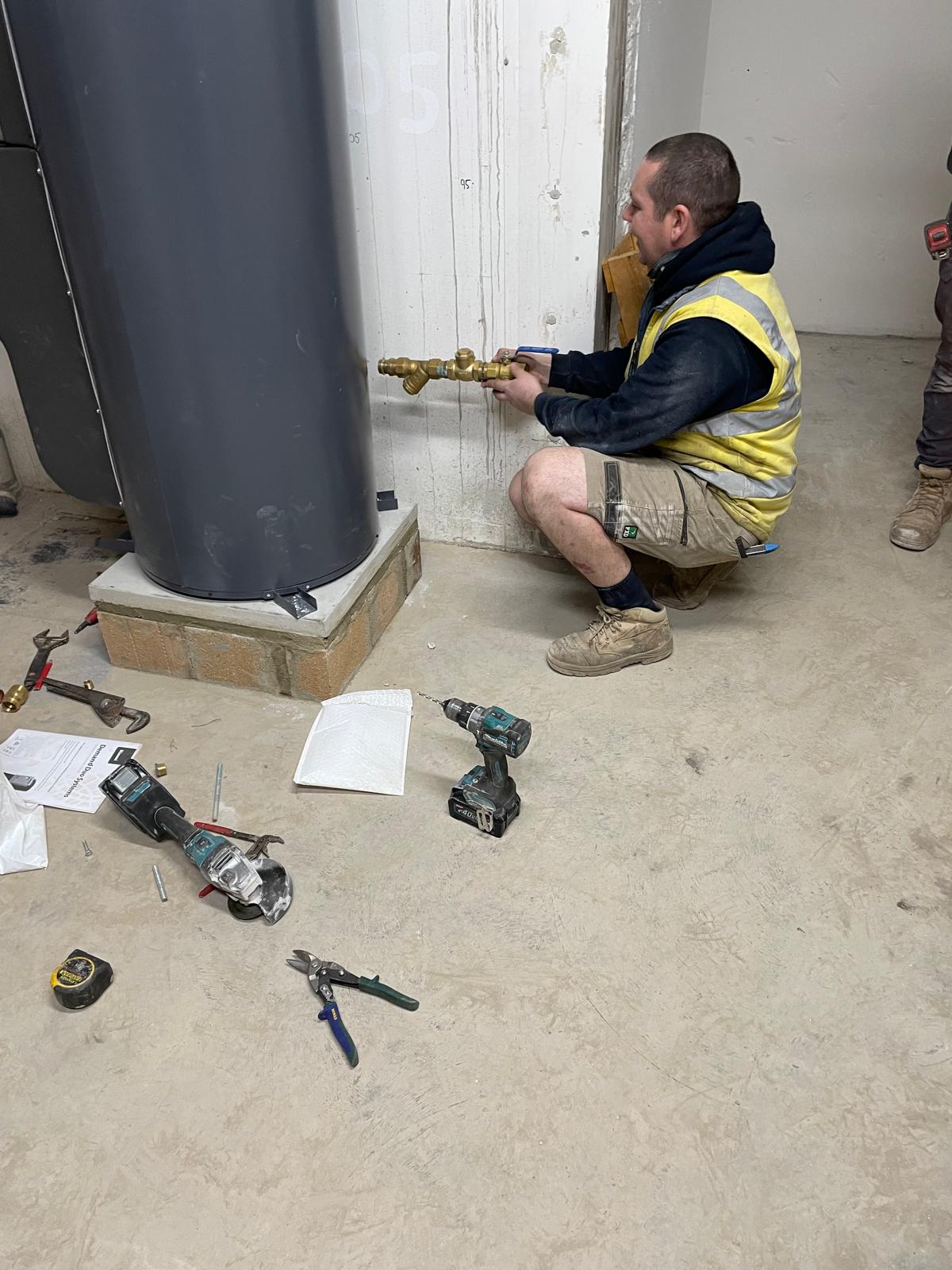A worker in a yellow vest kneels, installing plumbing near a cylindrical structure in Surry Hills. Tools scattered nearby.