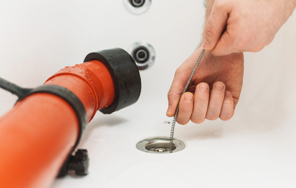 Plumber using a drain snake to clear a blocked bathtub drain with drain cleaning equipment nearby.